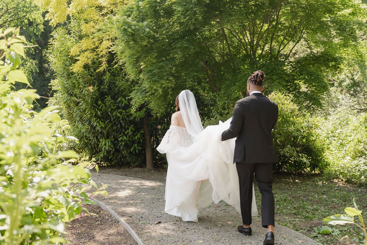 A couple in wedding attire walks along a tree-lined path surrounded by lush green foliage in a garden setting.
