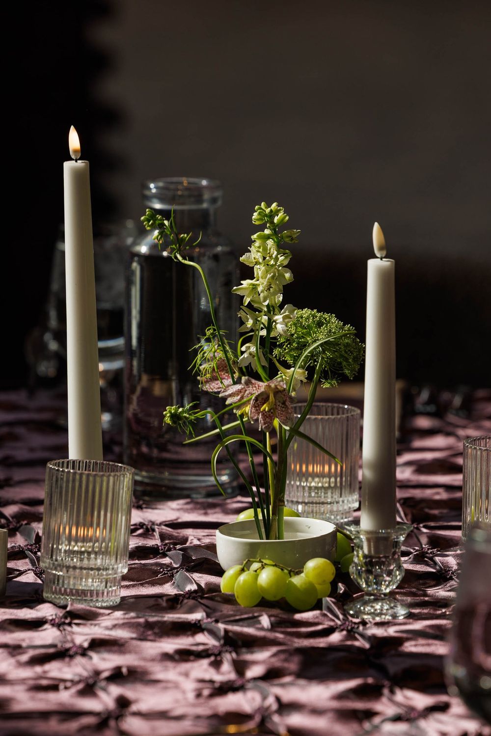 Elegant table setting with white candles, green apples, and parsley sprigs on a dark purple tablecloth in moody lighting.