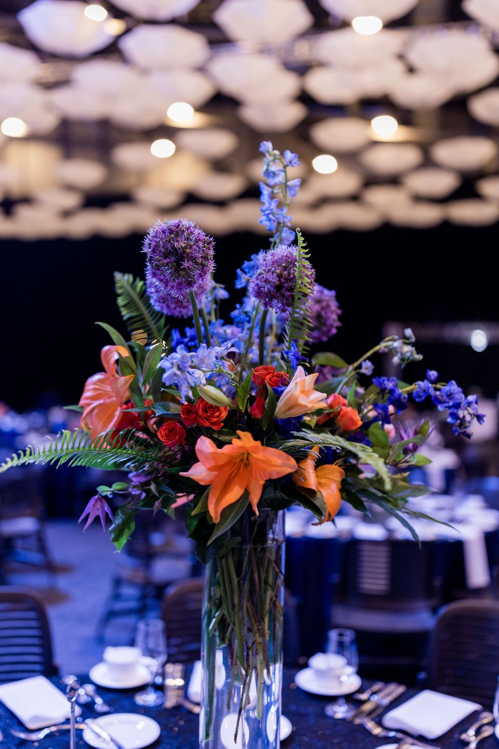 Elegant floral centerpieces with orange lilies and purple flowers in glass vases on formal dining tables with bokeh lighting.
