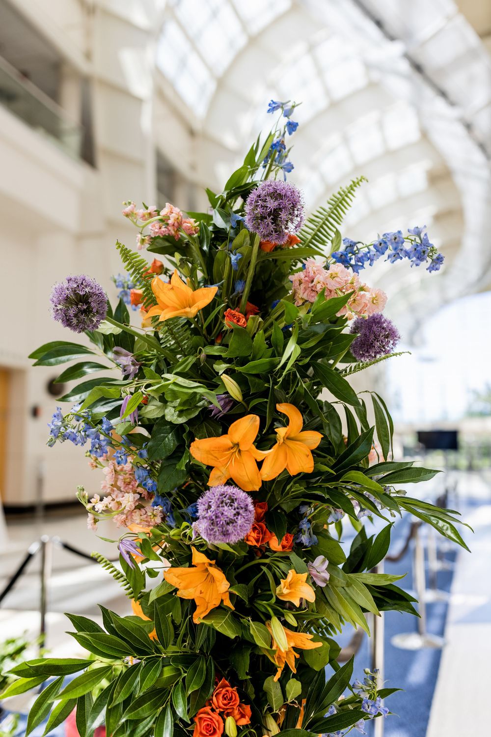 Vibrant floral arrangement featuring orange lilies, purple allium, and green foliage in a bright indoor setting.