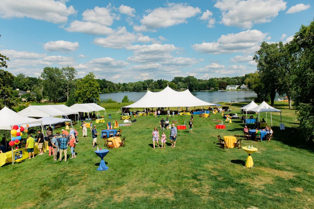 Outdoor summer event with white tents and colorful activities set up on a grassy lawn next to a lake on a sunny day.