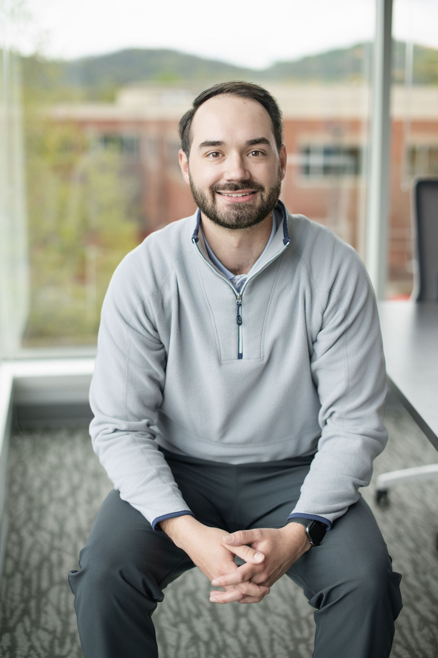 A person in a gray quarter-zip sweater sits casually on a bench with blurred background scenery.