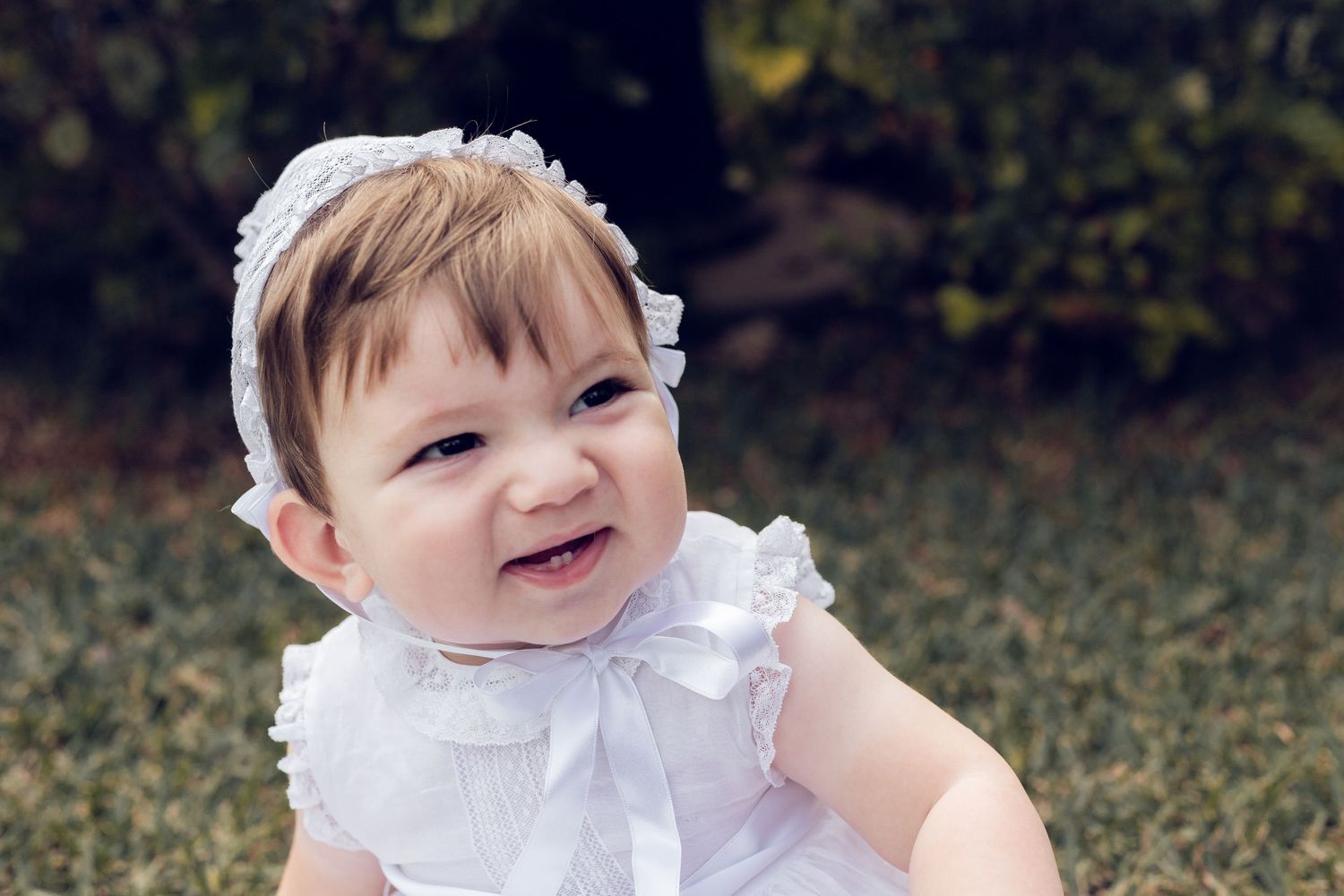 A baby in a white ruffled dress sitting and smiling on grass outdoors.
