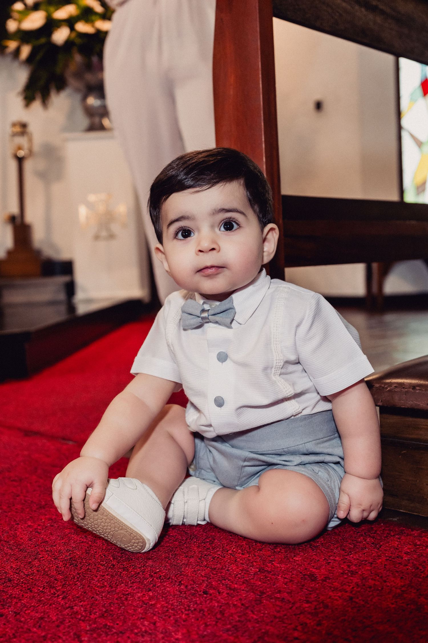 A baby in a white shirt and gray shorts sits on a vibrant red carpet in a series of candid poses.