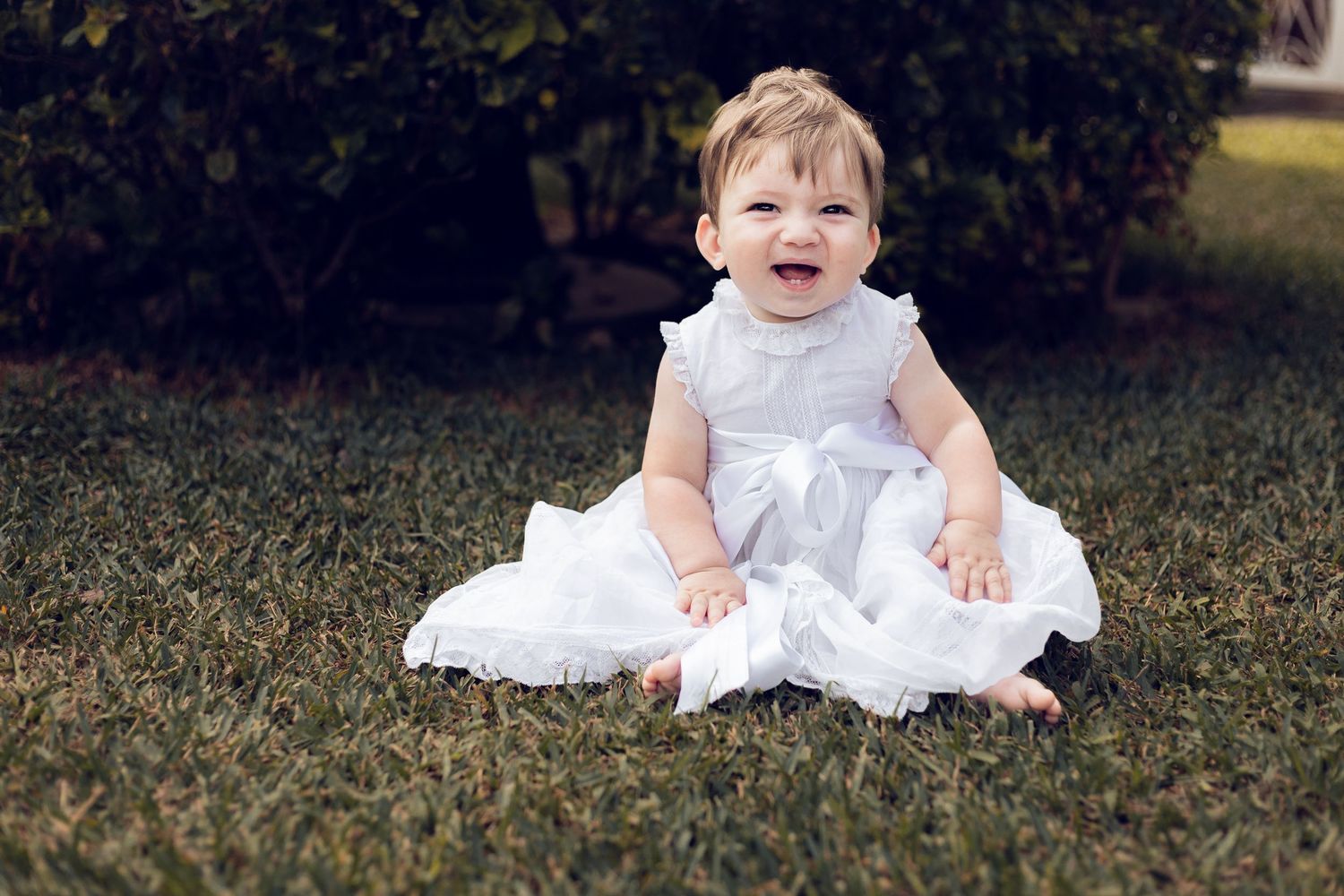 A baby in a white dress sits on grass while laughing and smiling during an outdoor photo session.
