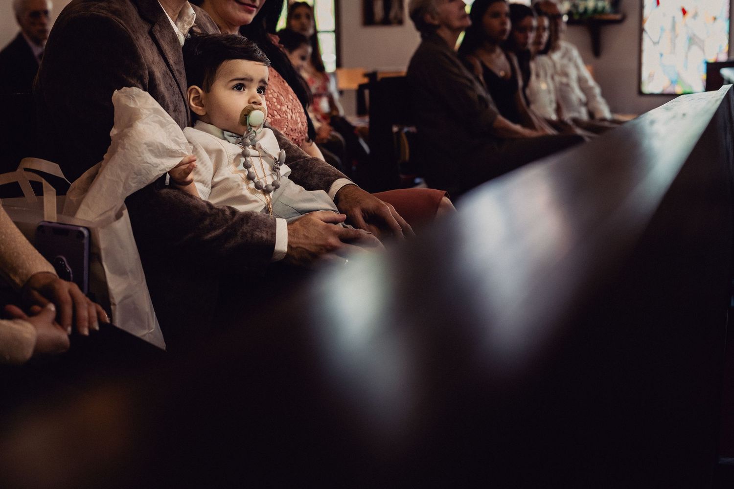 Young child in white attire sits at dark table during indoor gathering.