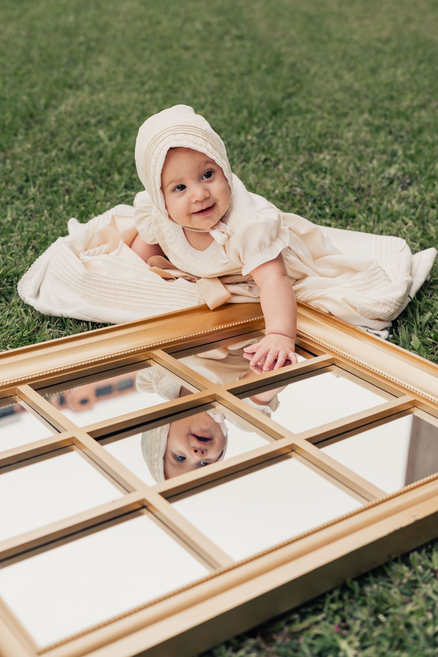 Baby in vintage dress lying next to decorative gold mirror frame on grass.