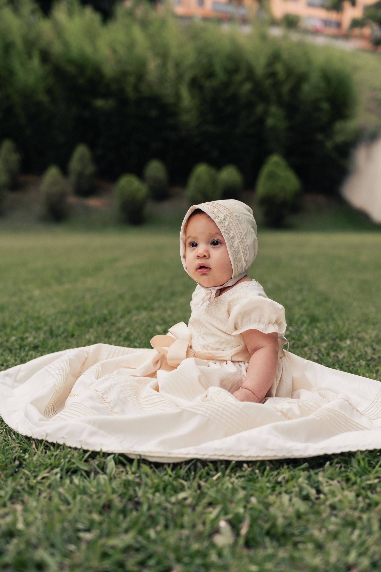 Baby wearing cream baptismal gown and bonnet resting on grass outdoors.