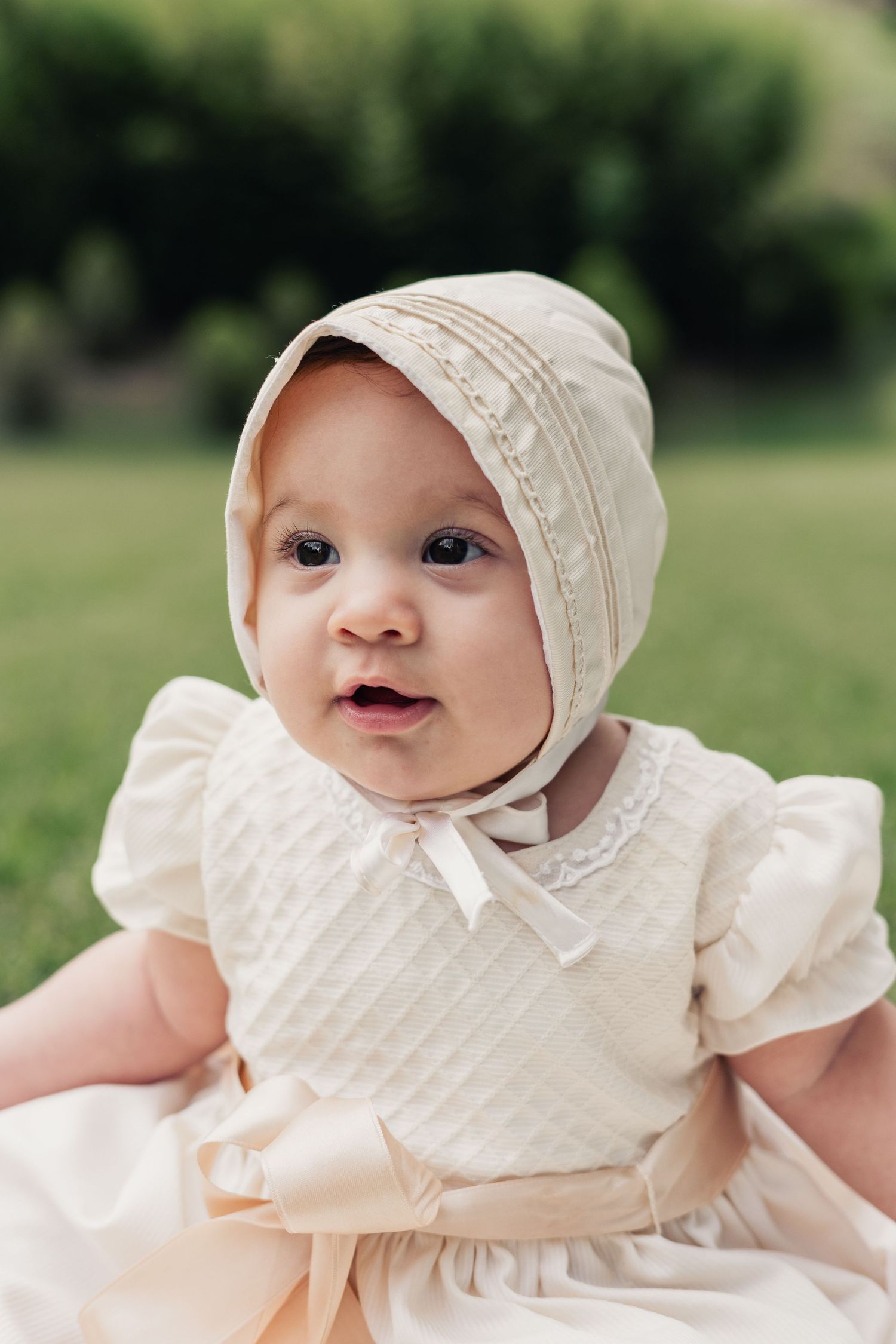 Close-up of baby in cream christening outfit with bonnet against blurred green background.