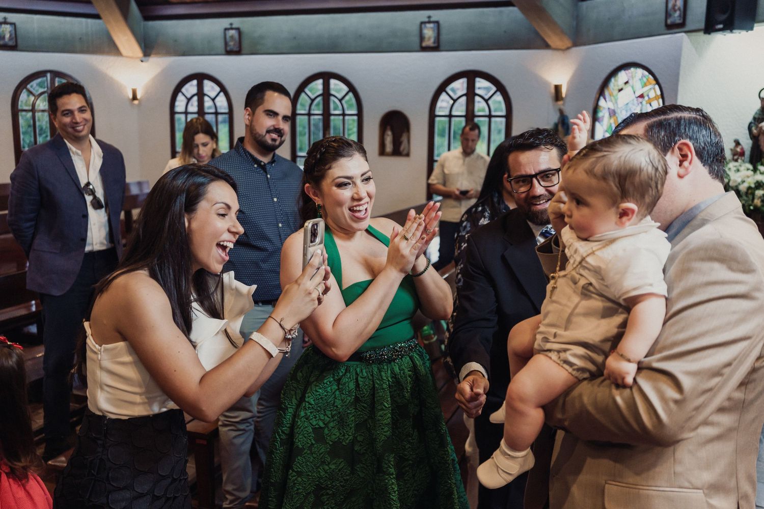 A group of wedding guests in formal attire gather around a small baby at a reception venue with arched windows.