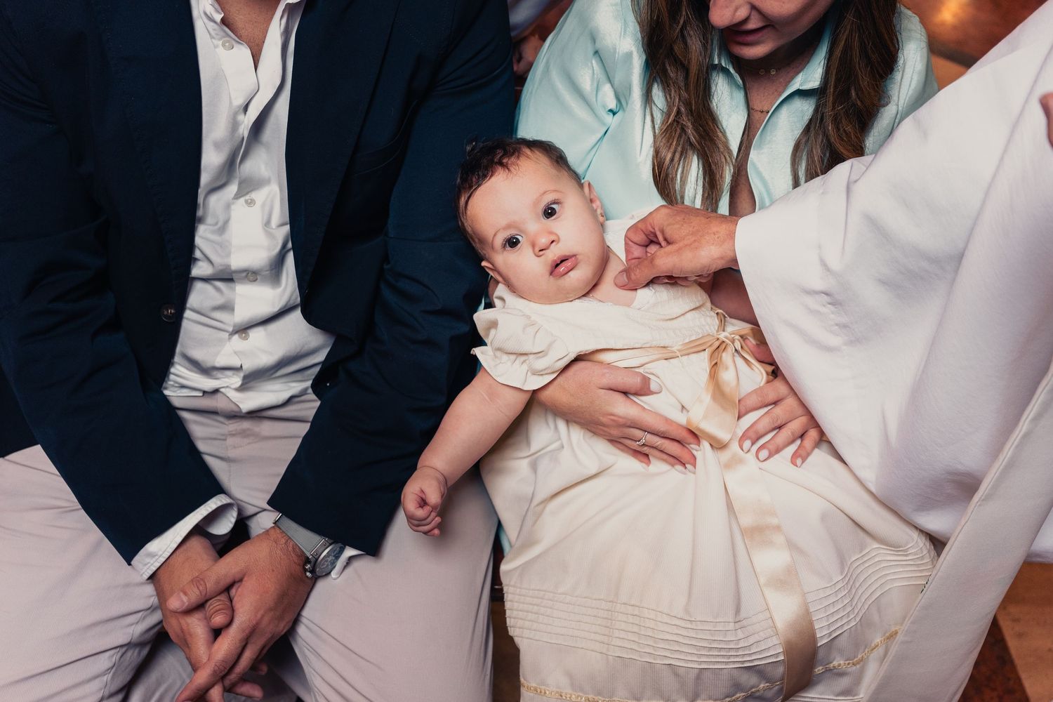 Family members in formal attire holding baby in white baptismal gown during ceremony.