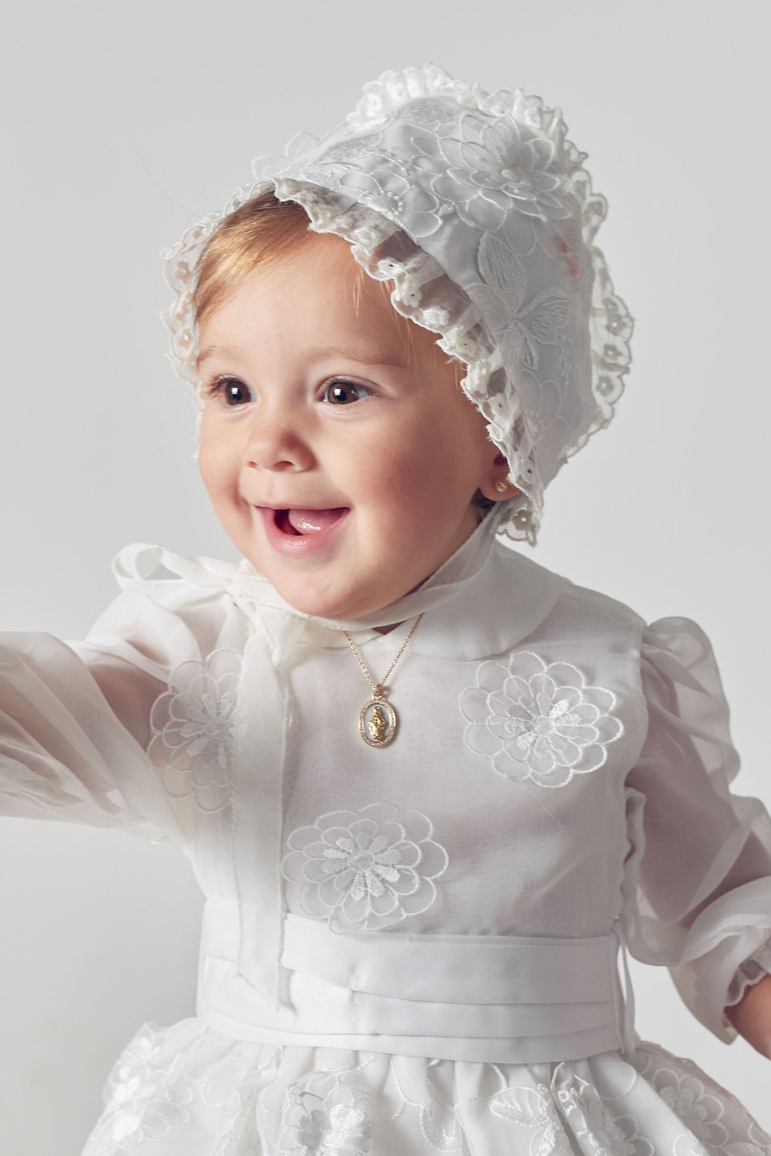 A baby wearing a traditional white christening gown and bonnet smiles while photographed in multiple poses.