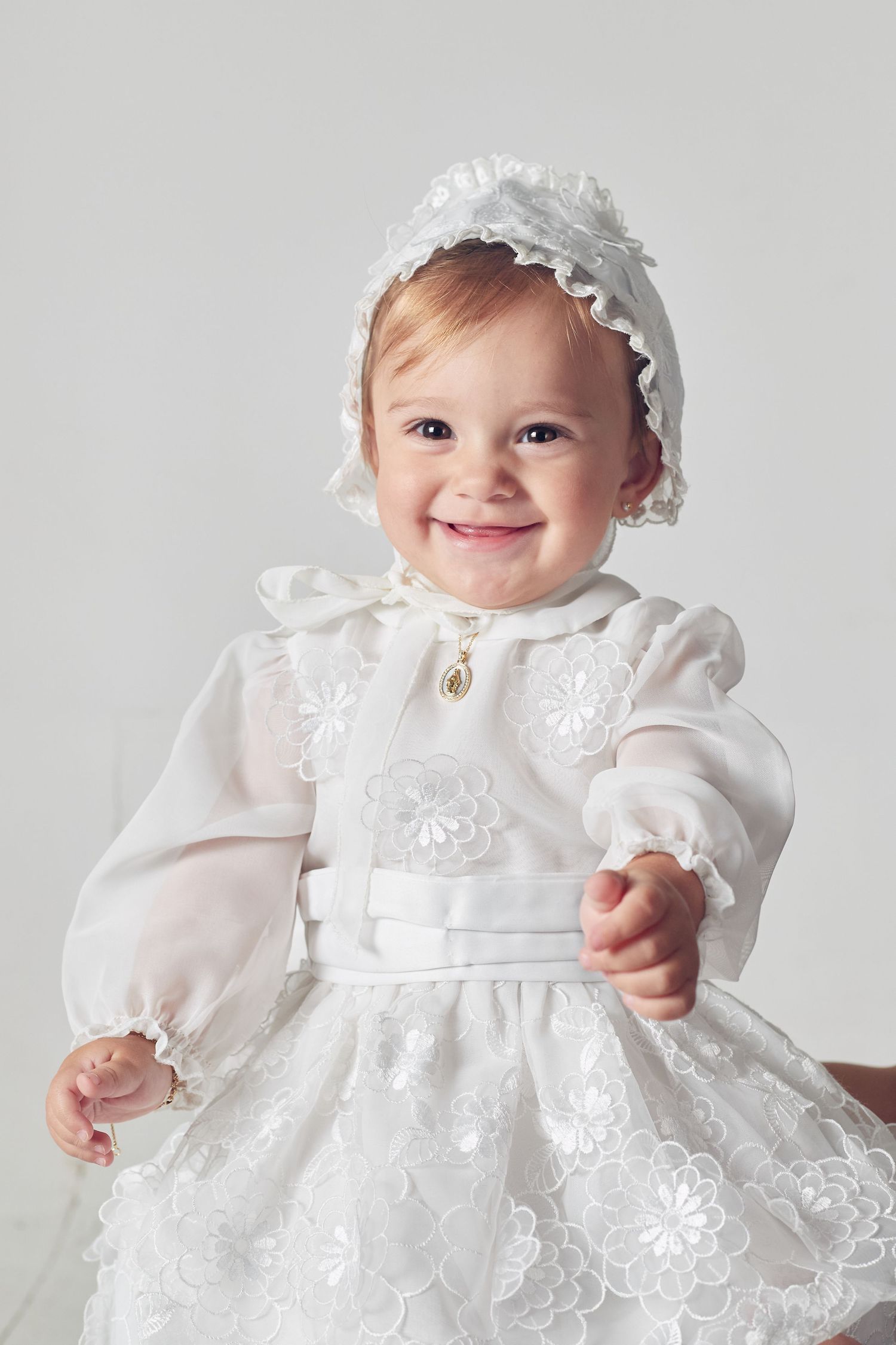 Baby wearing an elegant white christening gown with lace details and a delicate headpiece smiles at the camera.