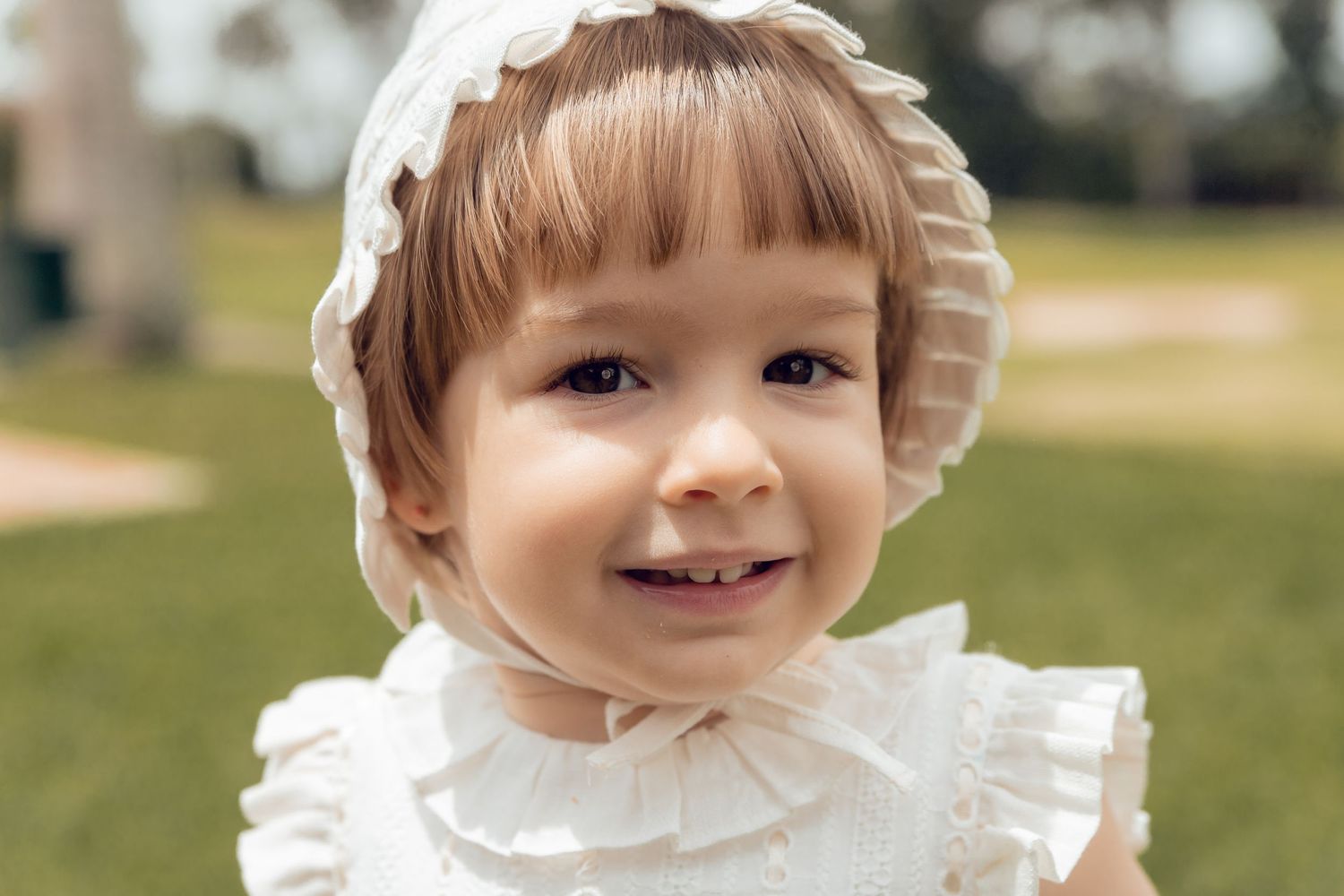 A child in a white ruffled dress smiles while playing outdoors on a sunny day.