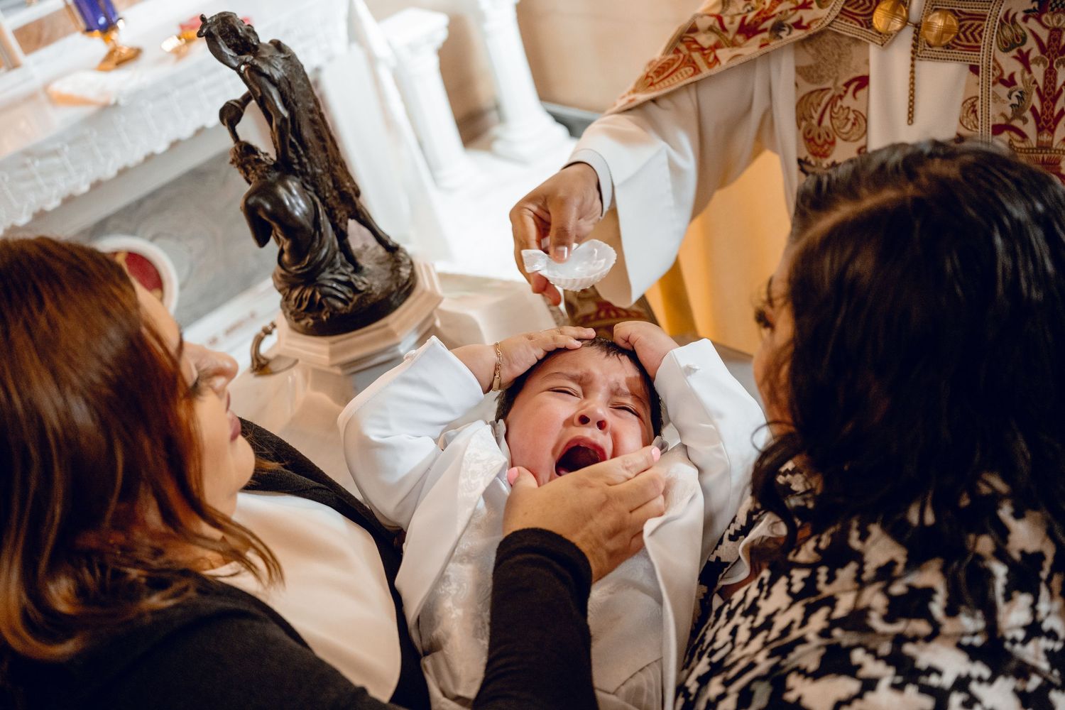 Baby in white clothing being held during baptism ceremony with holy water.