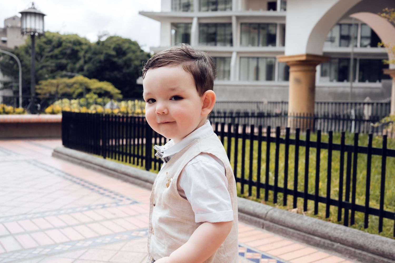 A young child in a white shirt sits on a decorative brick wall with buildings visible in the background.