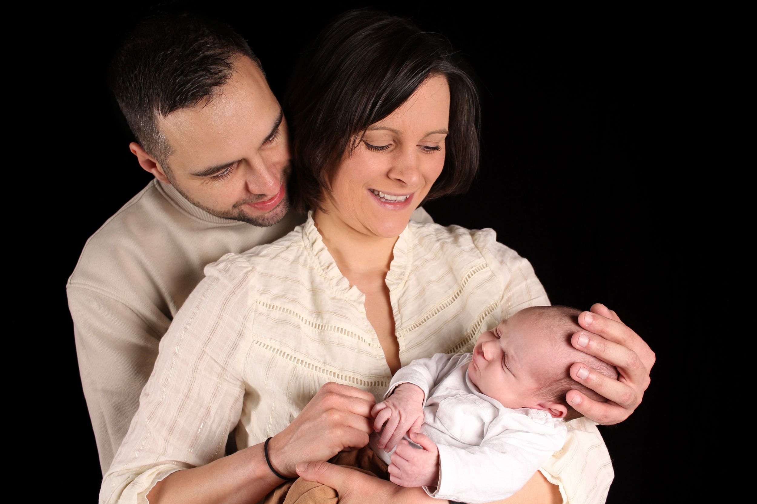 Séance photo naissance studio, jeunes parents tenant leur nouveau-né endormi dans leurs bras sur fond noir.
Lumeria Photo Montfort sur meu