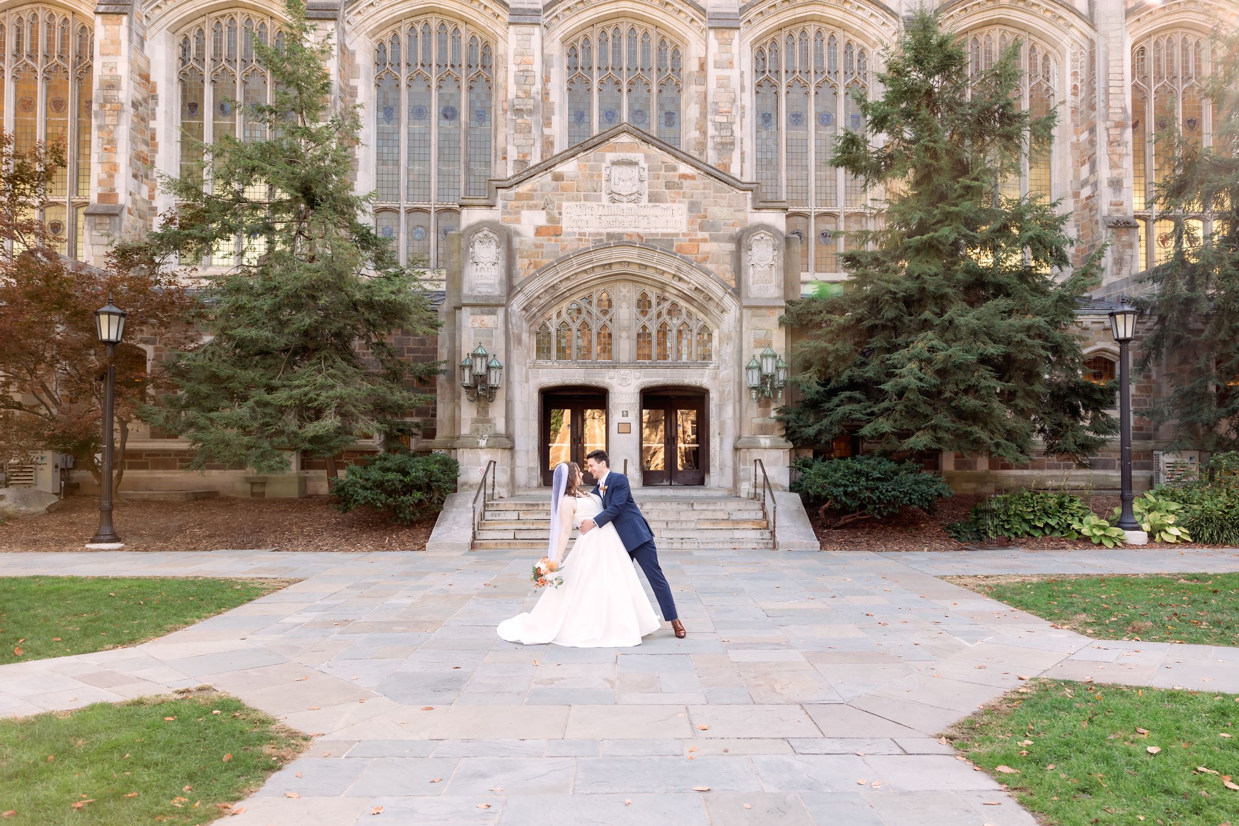 Elegant couple poses for wedding photos in front of a grand Gothic stone church entrance on a sunny autumn day.