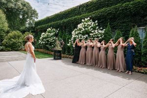 A wedding party in mauve bridesmaid dresses stands in line against an ivy-covered wall outdoors.