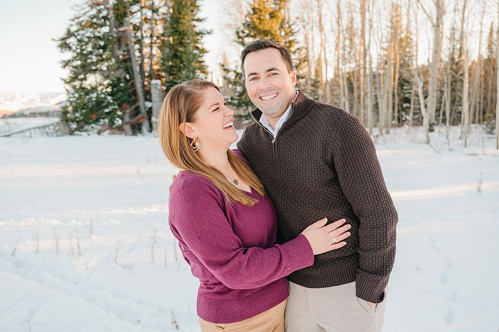 Couple embracing in a snowy winter setting with white birch trees and evergreens behind them.