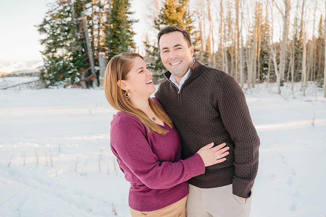 Couple embracing in a snowy winter setting with white birch trees and evergreens behind them.