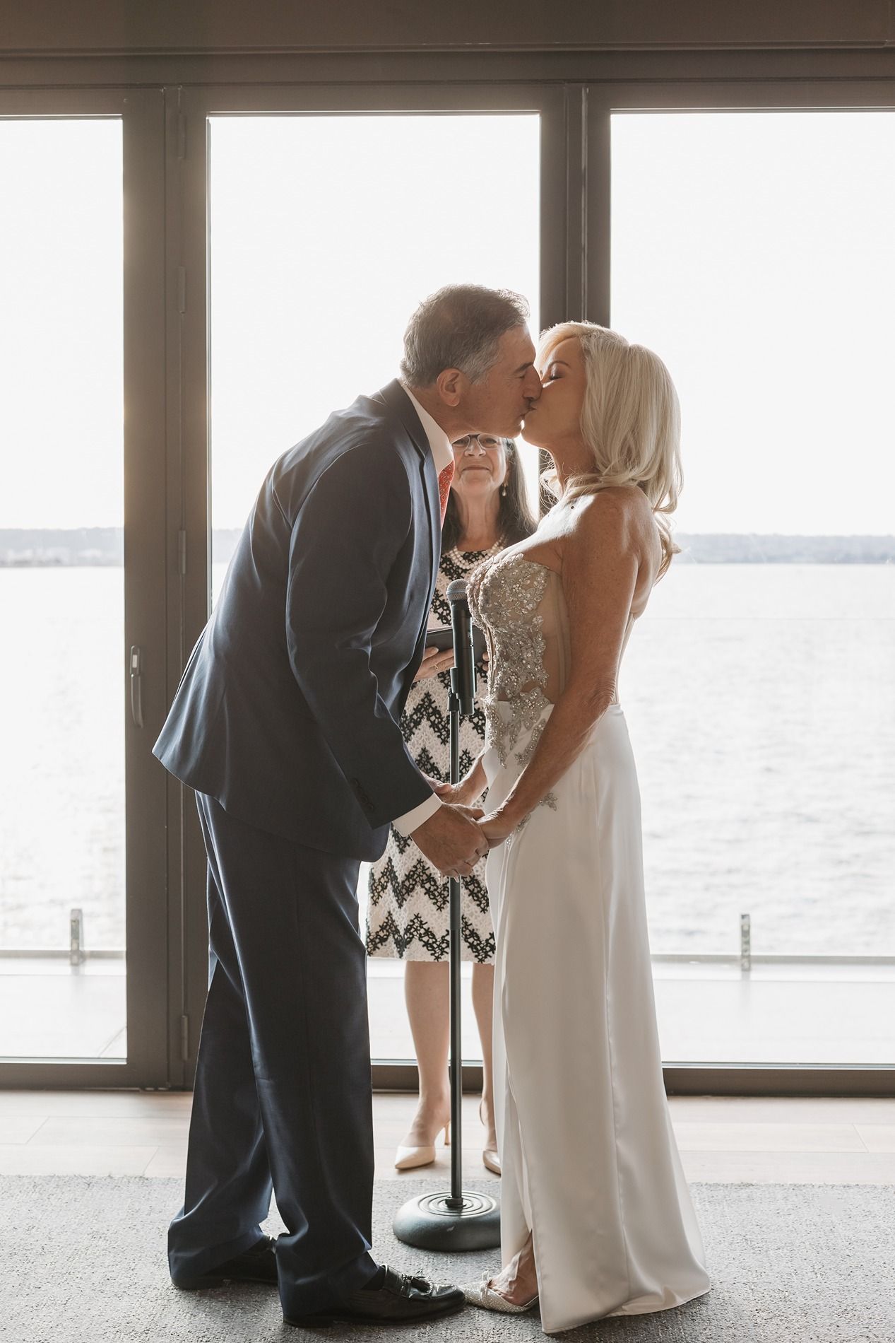 A romantic wedding moment with a couple sharing a kiss by large windows overlooking a waterfront view.