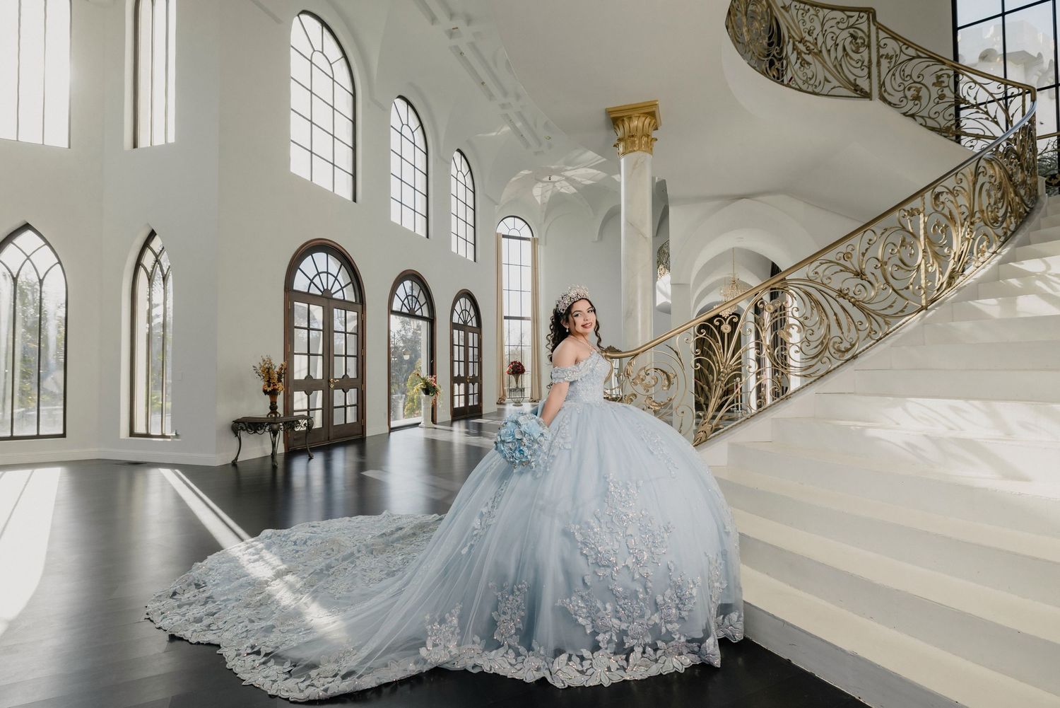 Grand white ballroom interior with ornate staircase and arched windows.