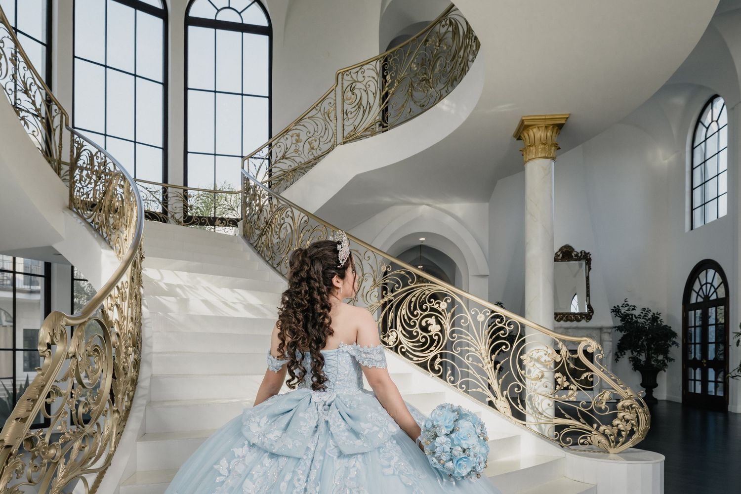 Elegant light blue ballgown on grand marble staircase with ornate gold railings and tall arched windows.