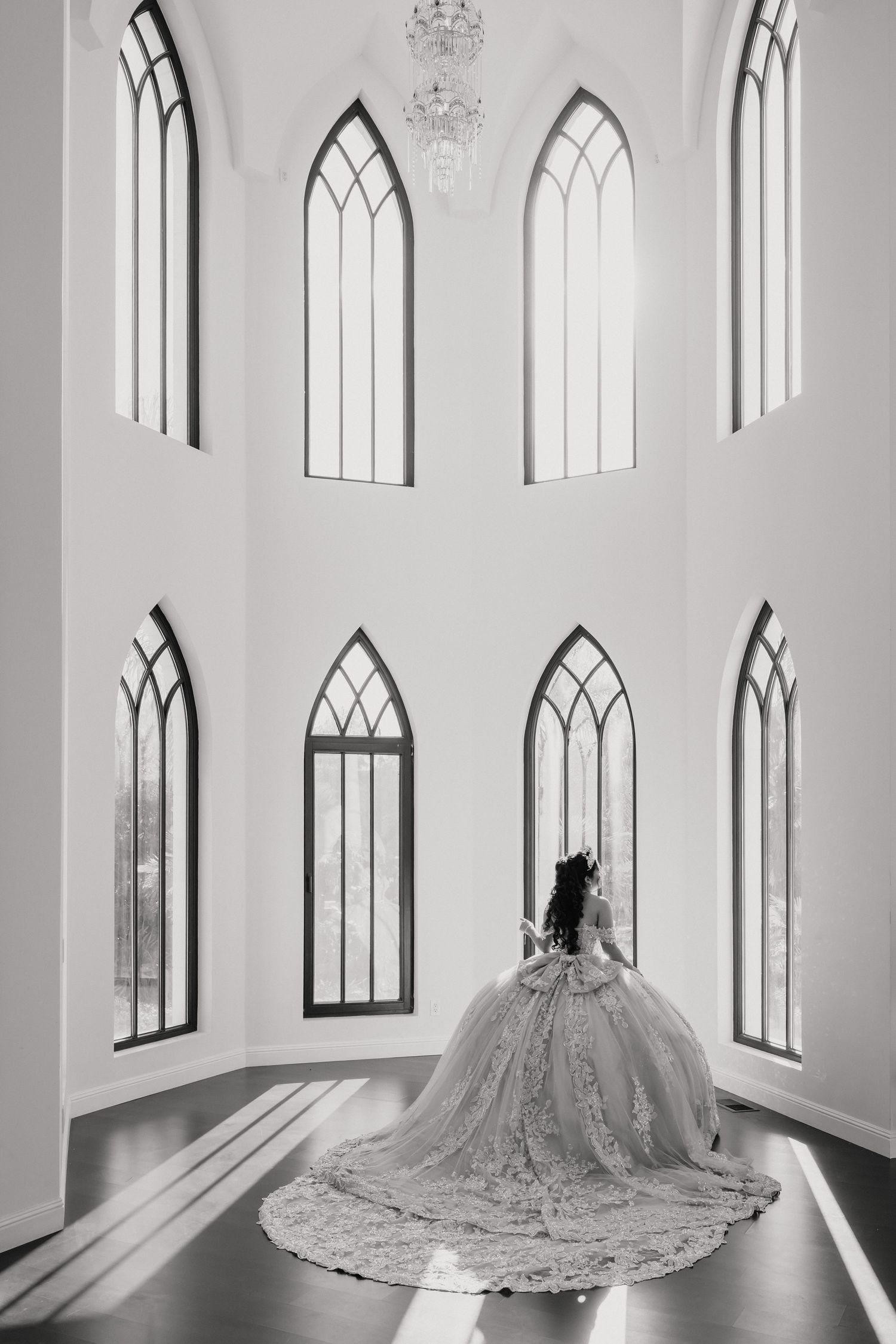A dramatic black and white photo of a ballgown with a flowing train in an elegant chapel room with gothic windows.