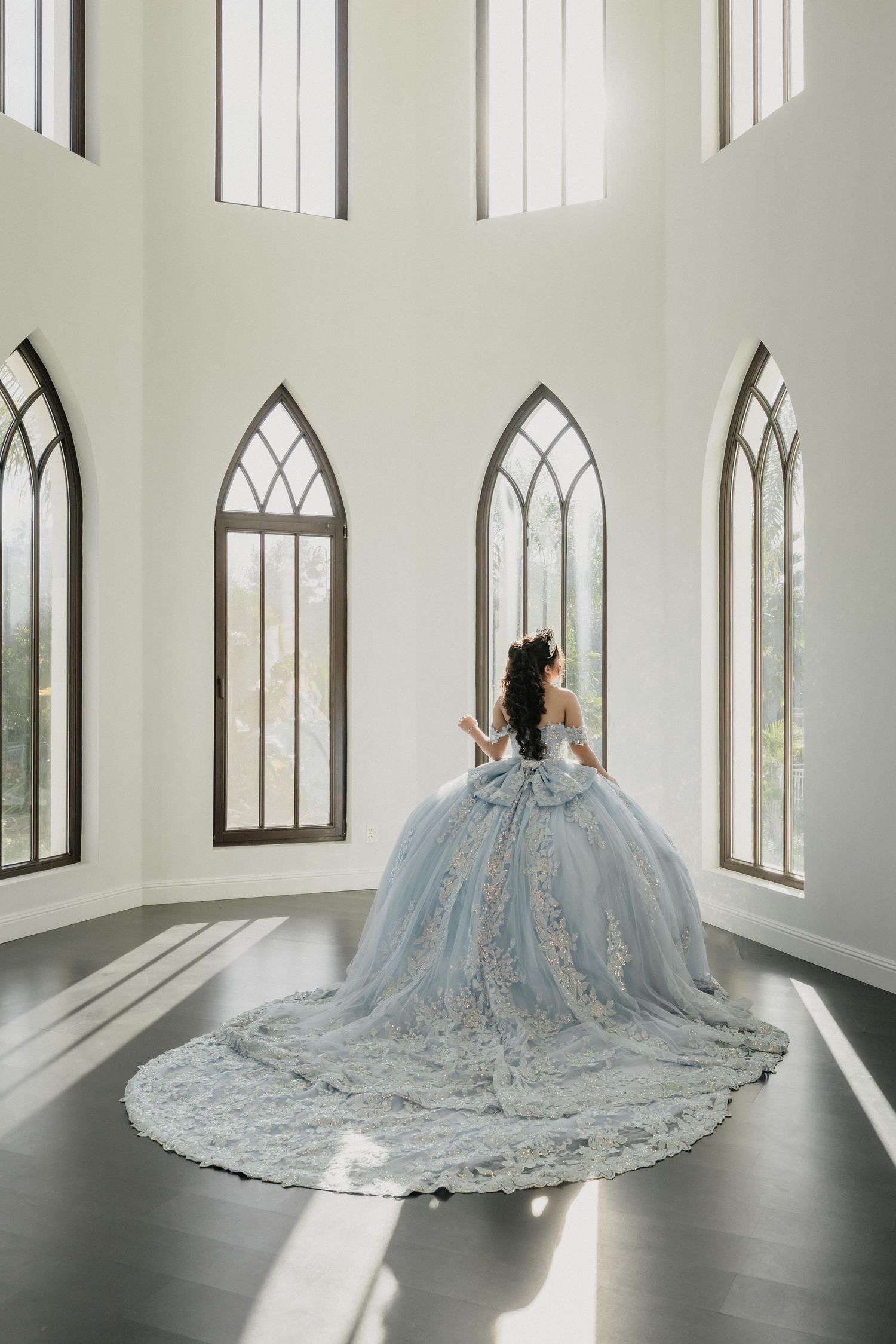 A person in a flowing light blue ball gown with intricate details stands in an elegant white chapel room with arched windows.