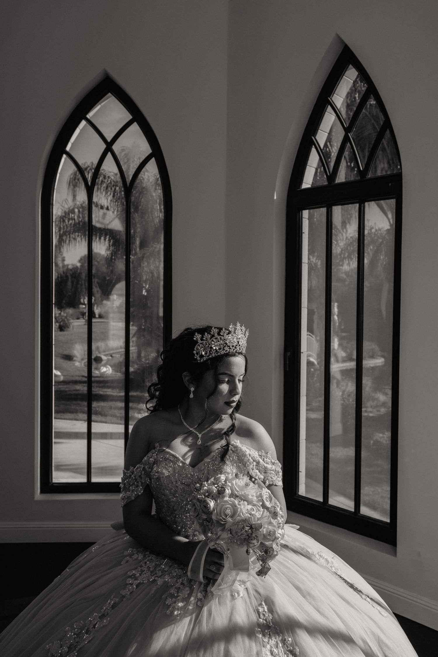 Dramatic black and white portrait showcasing an ornate ball gown and crown against gothic windows.