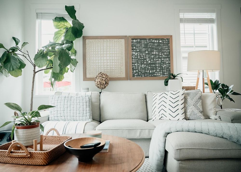 Modern living room with gray sofa, wooden coffee table, decorative pillows and potted plants by sunlit windows.