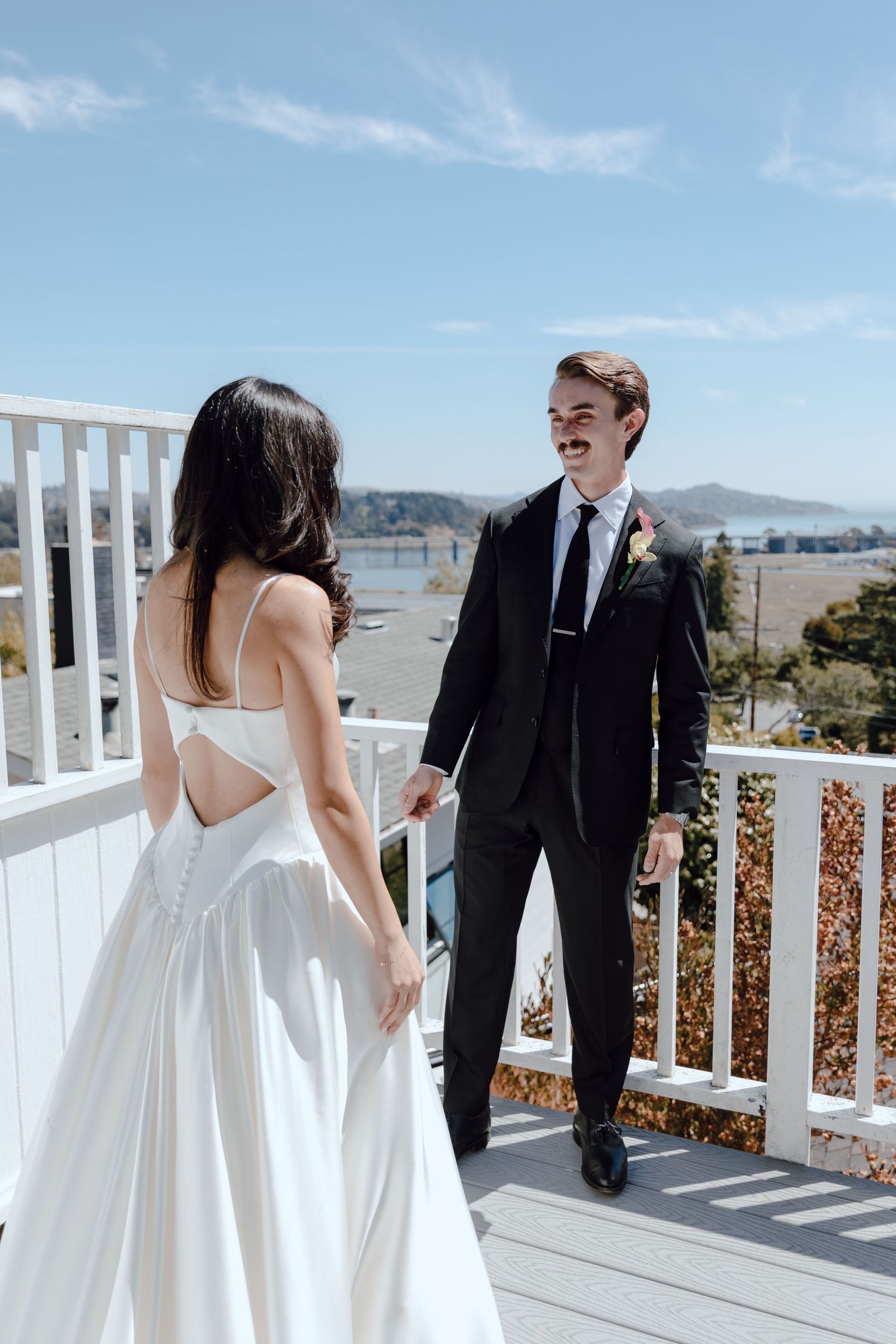 A couple shares their first look moment on a white deck balcony overlooking a scenic mountain and water view.
