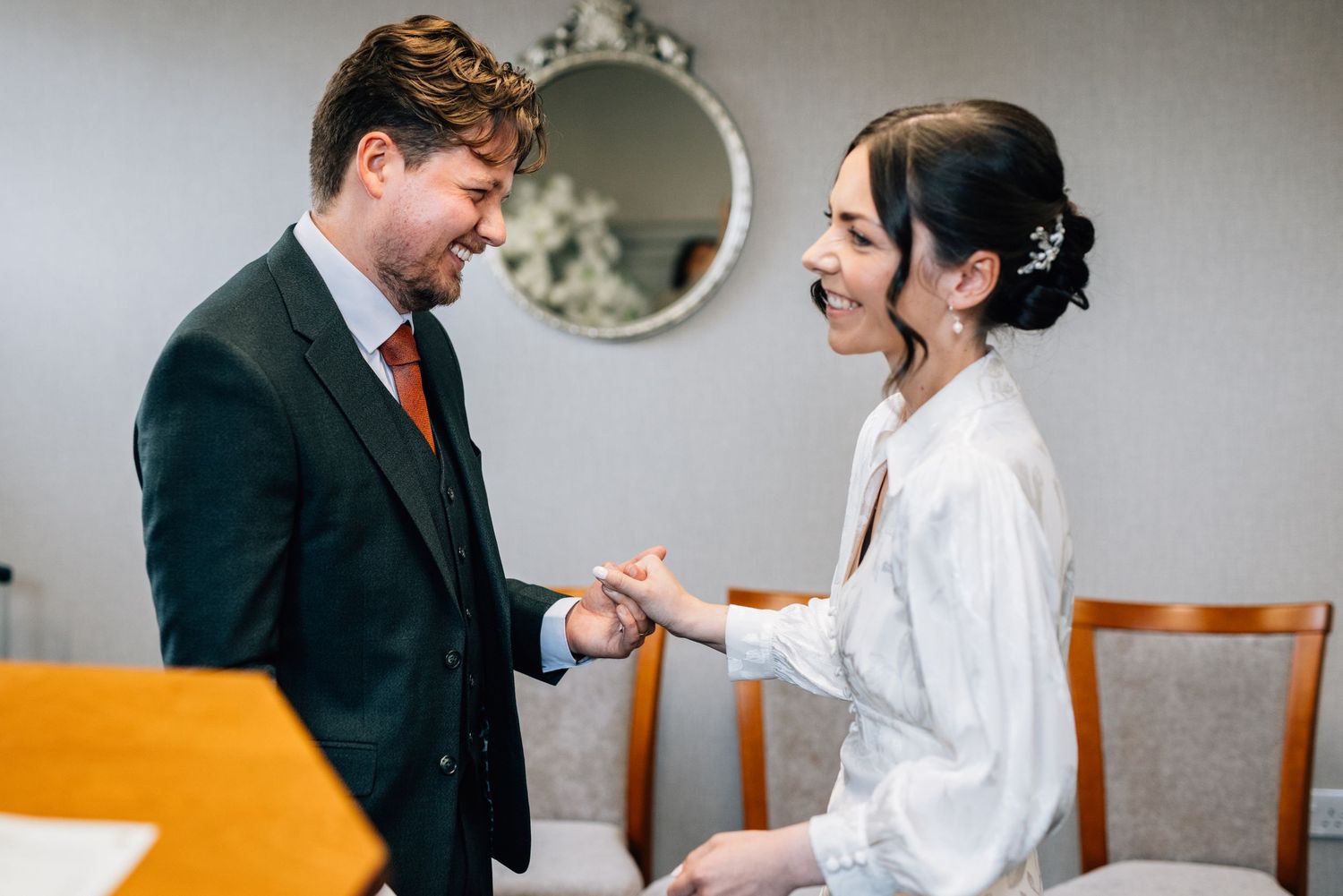 A couple in formal attire sits at a table together smiling and holding hands in an intimate moment.
