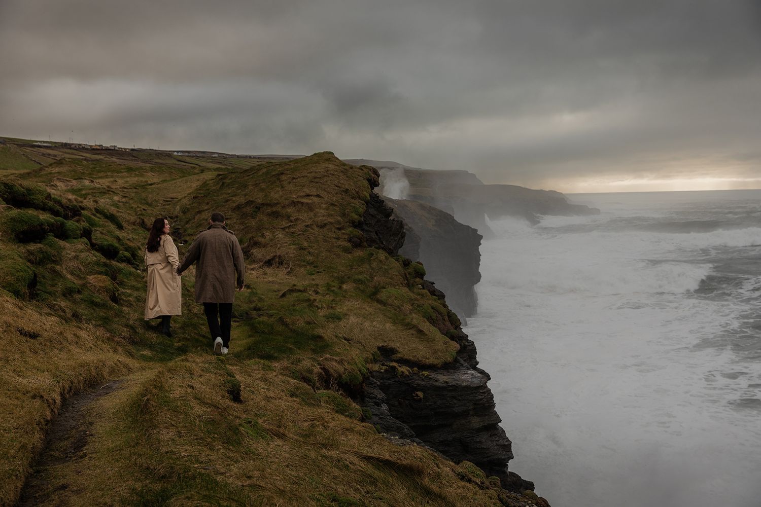 Couple walking on Cliffs of Moher during stormy coastal engagement session at golden hour