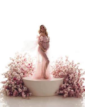 A dreamy maternity photoshoot featuring pink florals and a feminine silhouette in a flowing dress against white backdrop.