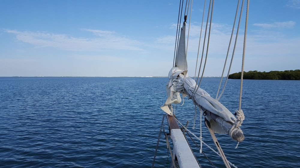 The bow of a sailboat points toward calm blue water and a distant shoreline, capturing the peaceful rhythm of life on the water.