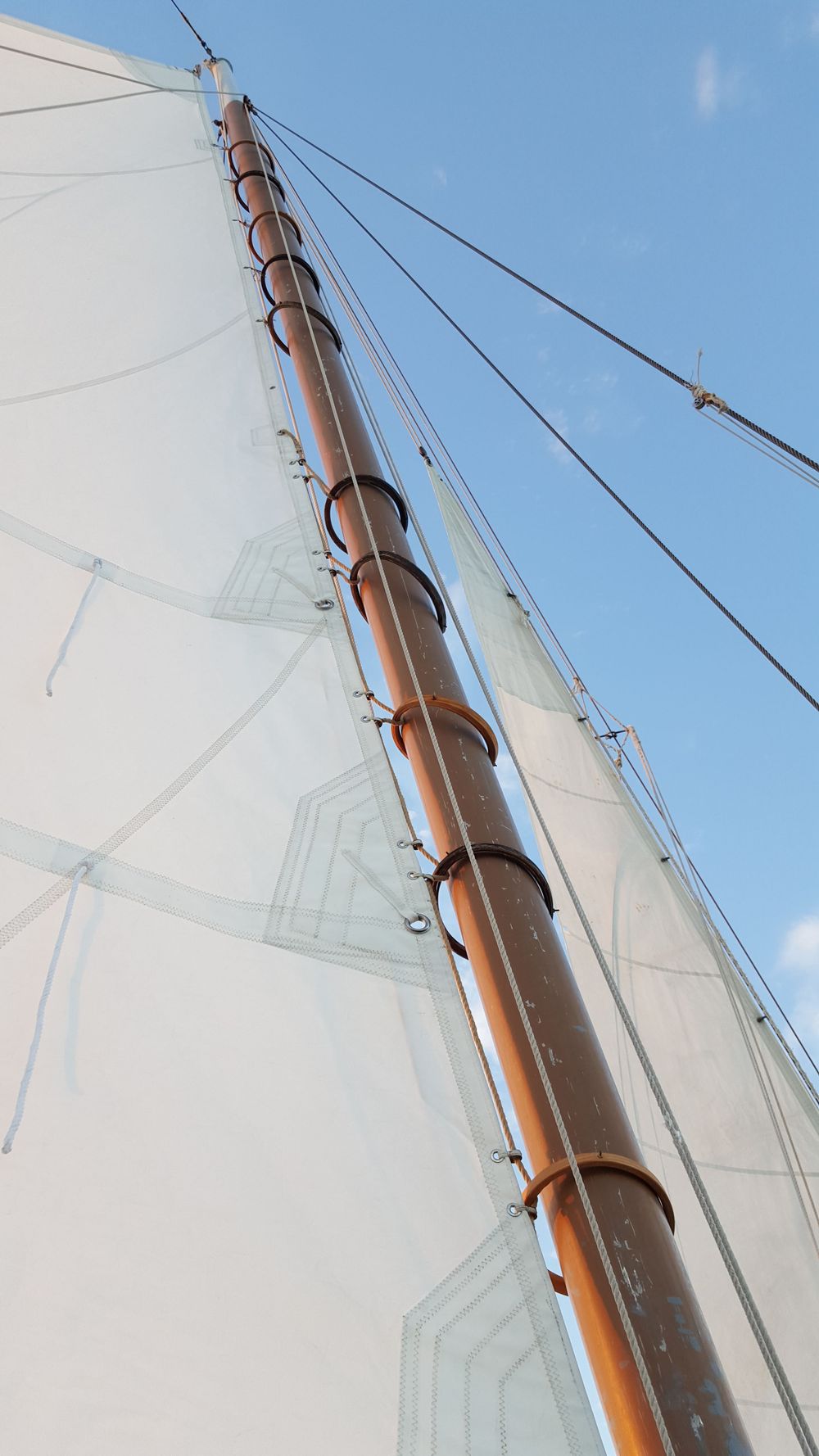 A sailboat mast rises into a clear blue sky, with crisp white sails and detailed rigging lines creating a clean, nautical composition.