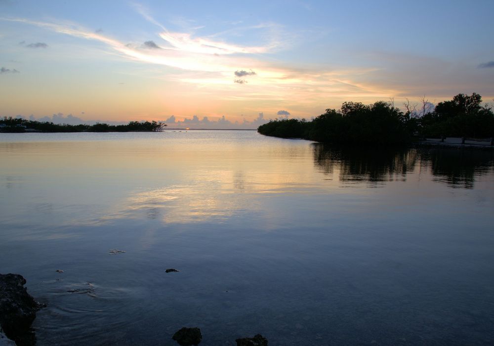 A warm sunset glows across a calm coastal waterway, with soft orange and yellow tones reflecting on the surface.