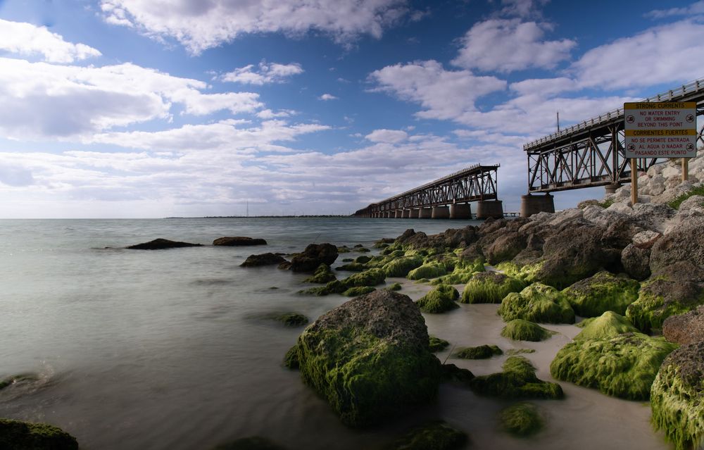 A long coastal railway bridge extends over calm water as green‑covered rocks line the shoreline.