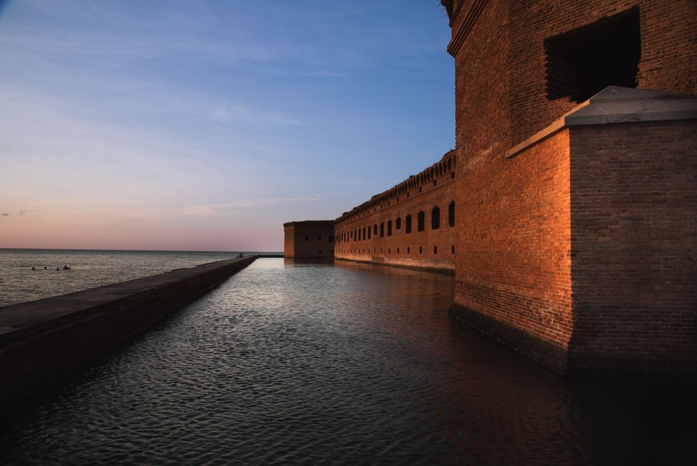 A historic brick fort glows in warm golden‑hour light as it stretches along the water’s edge. A stone walkway leads toward the horizon.