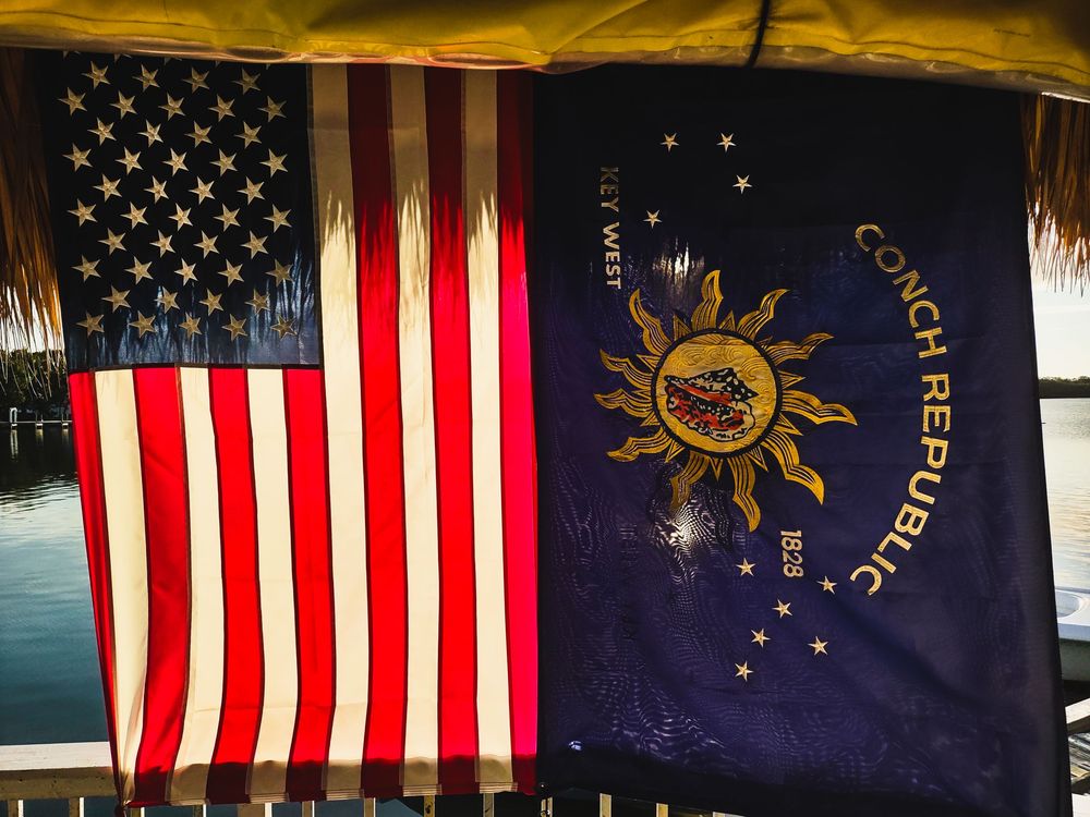 The U.S. flag and the Conch Republic flag fly side by side near a waterfront.