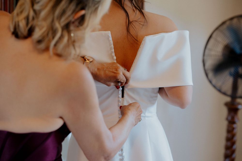 Helping to fasten the back of an elegant white wedding dress with off-shoulder detail.