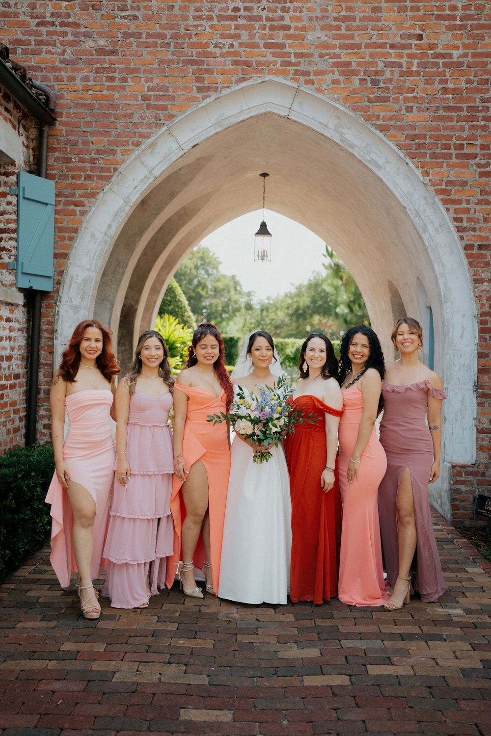 Bridesmaids in coral and pink dresses pose under a brick archway during a wedding photoshoot.