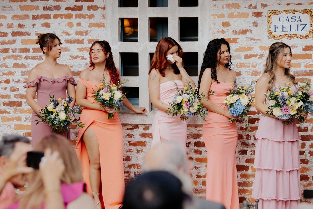 Bridesmaids in peach and pink dresses stand in a row holding floral bouquets against a brick wall.