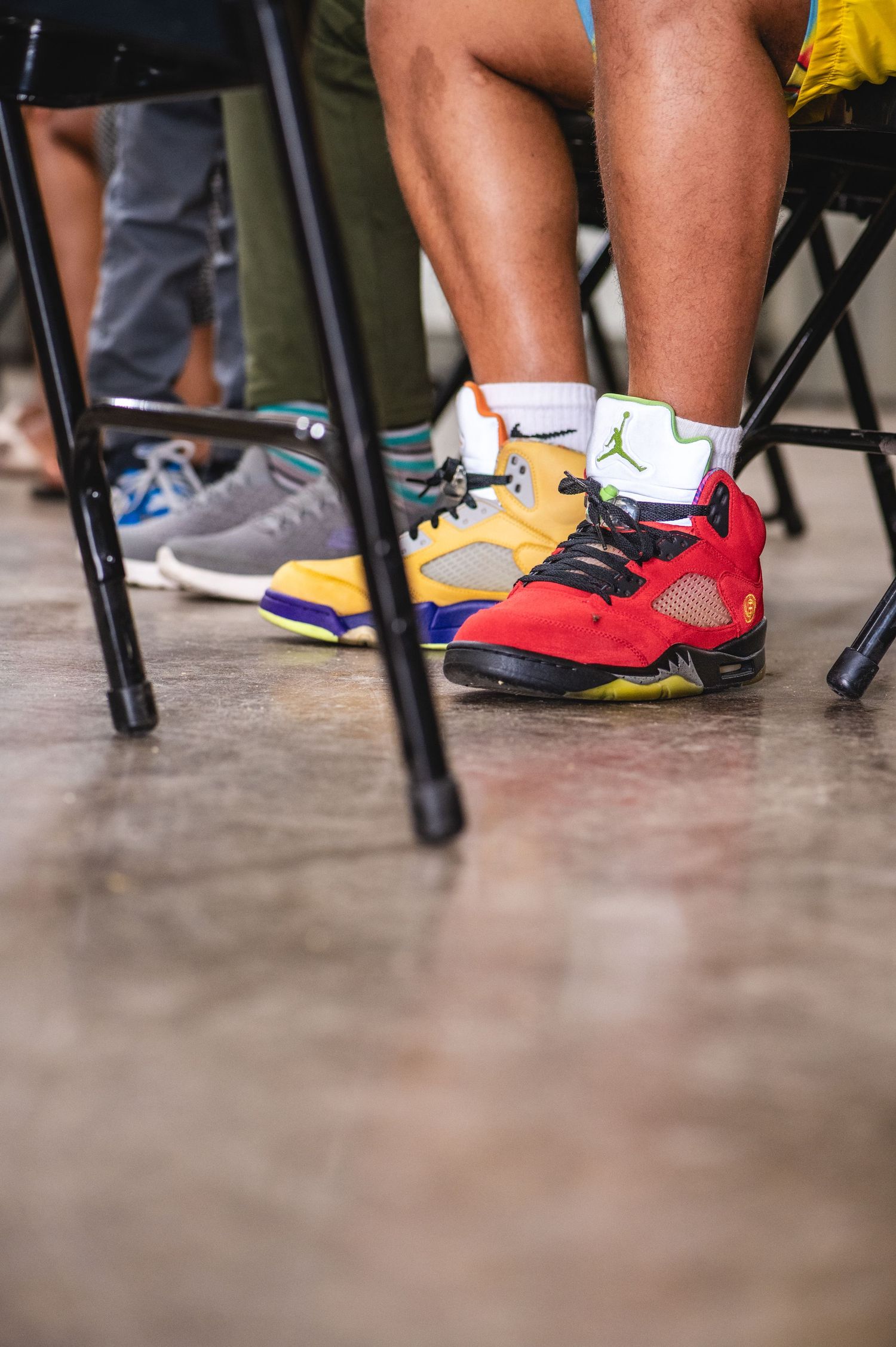 People wearing colorful sneakers including Air Jordans and Nike shoes sitting on chairs with black metal legs on concrete floor.