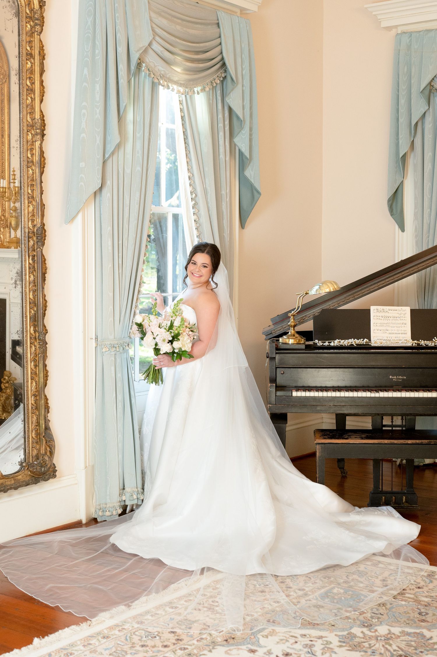 A flowing white wedding gown with a cathedral-length train is displayed in an elegant room with a grand piano.