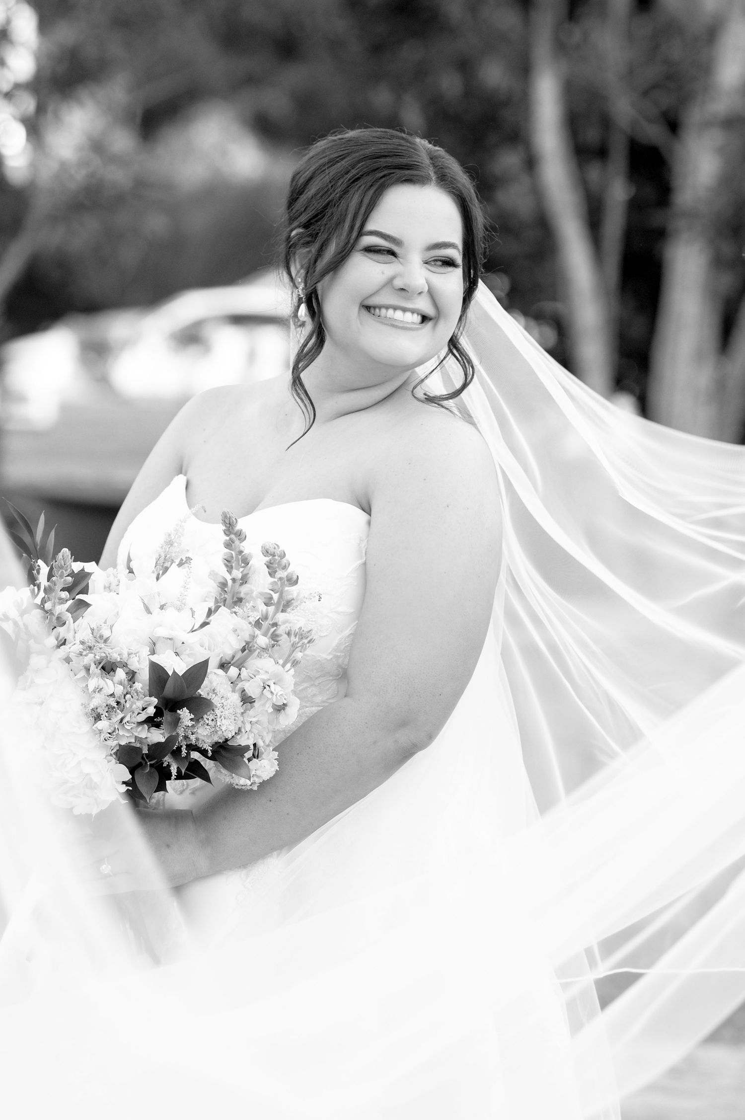 Black and white wedding photo showing joyful moment with flowing veil and delicate floral bouquet in natural setting.
