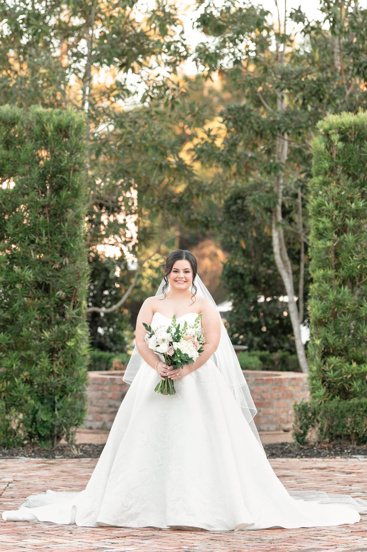 A bride in a flowing white wedding dress poses with her bouquet in a garden setting during golden hour.