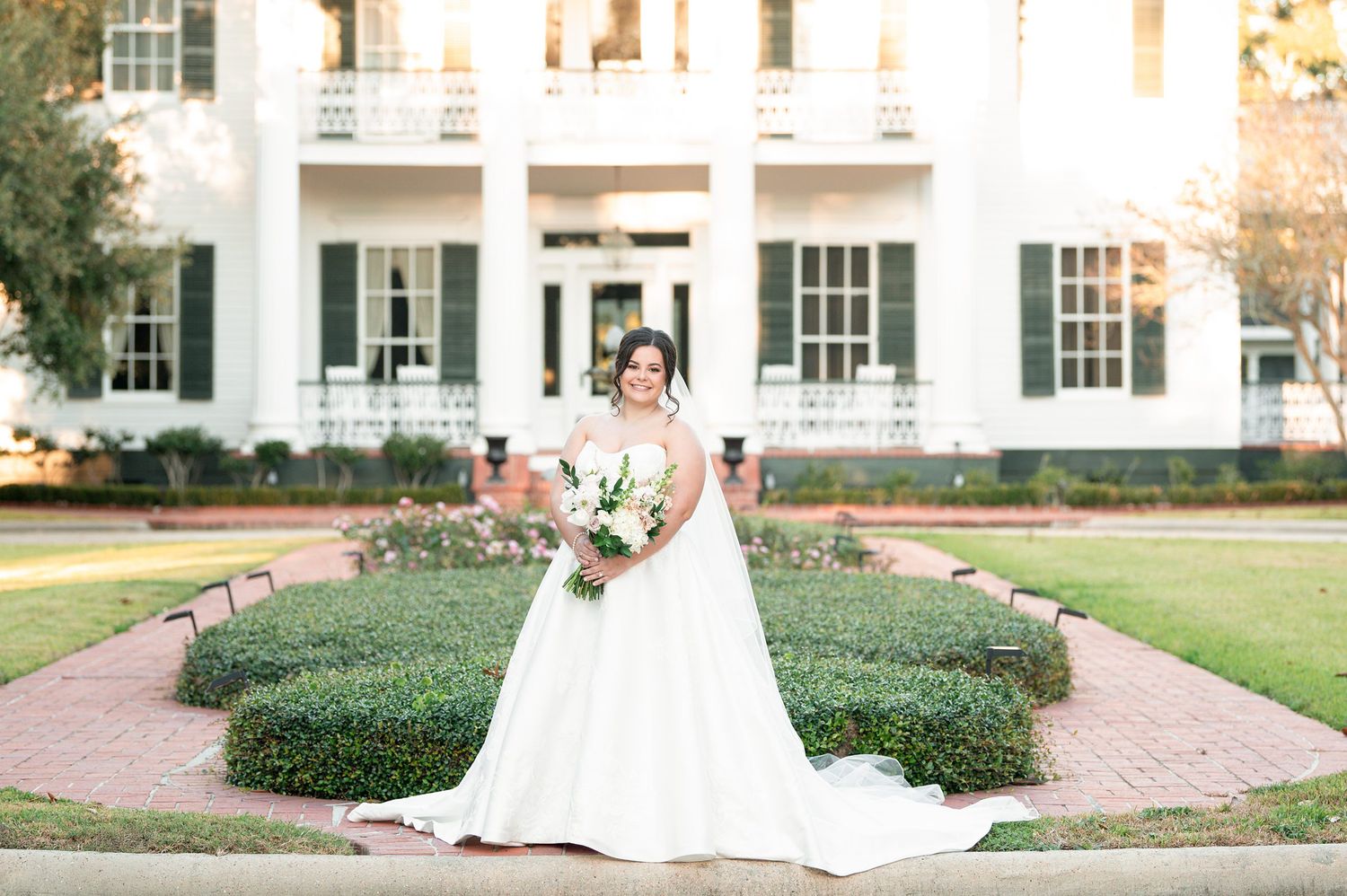 A wedding dress flows gracefully along a garden path lined with manicured hedges and historic architecture.